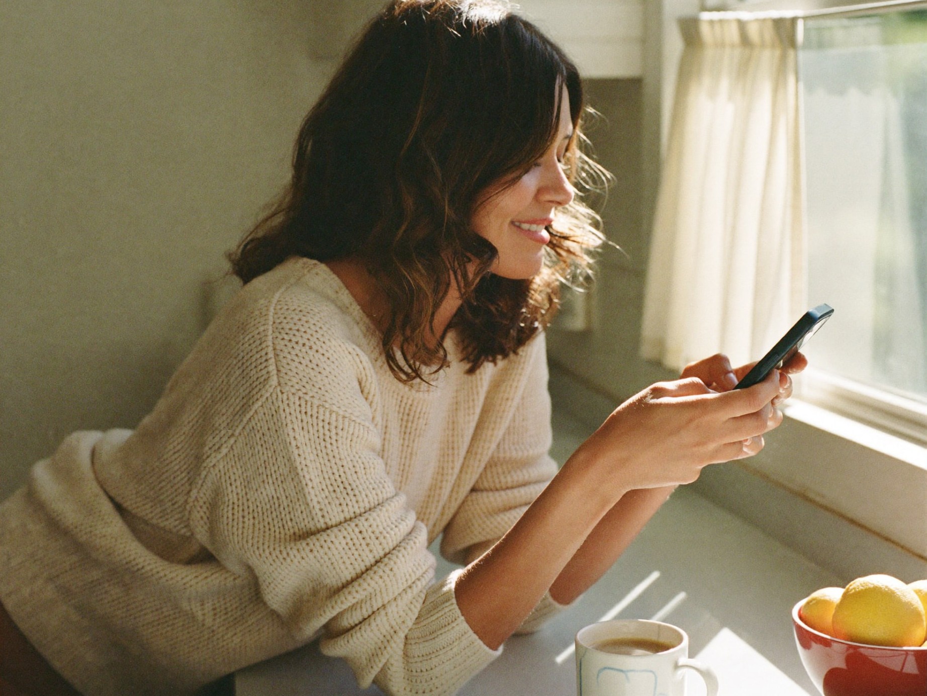 A smiling woman checking her phone at a sunlit kitchen counter